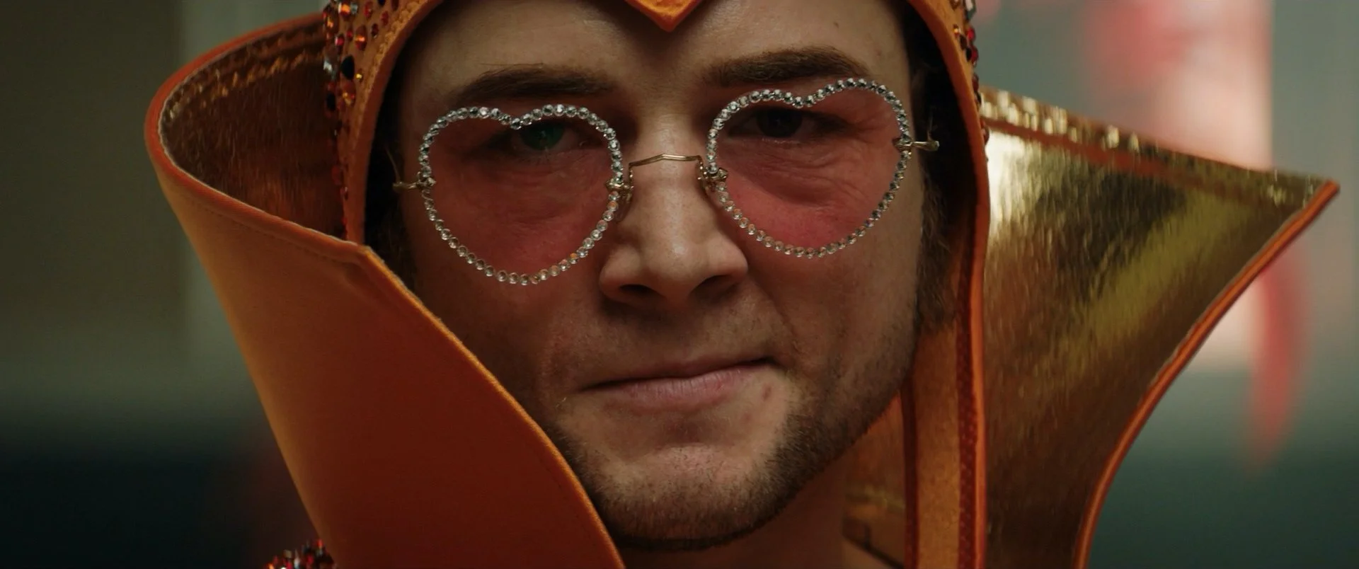 Close-up of a man wearing embellished heart-shaped sunglasses, an ornate orange headpiece, and a matching cape with reflective gold details.