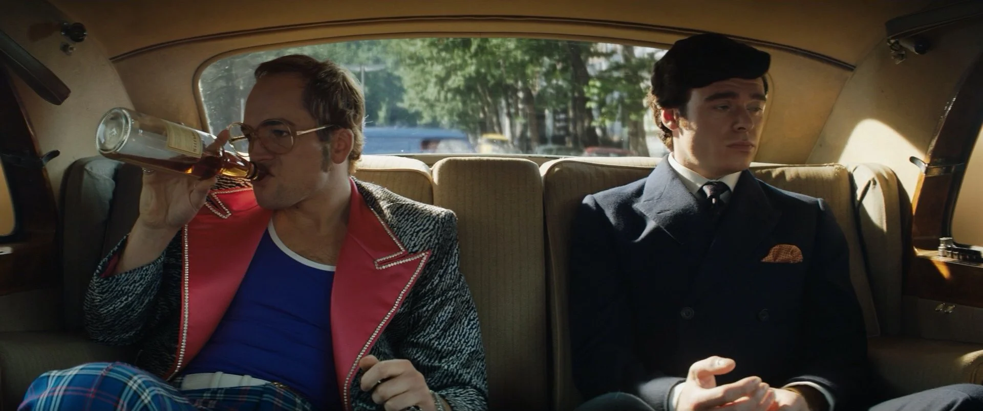 Two men sitting in the back of a car, one on the left drinking from a bottle, wearing glasses, a red and black jacket, and plaid pants, and the other on the right wearing a dark suit and tie, with a serious expression, during daytime with trees visible outside the window.