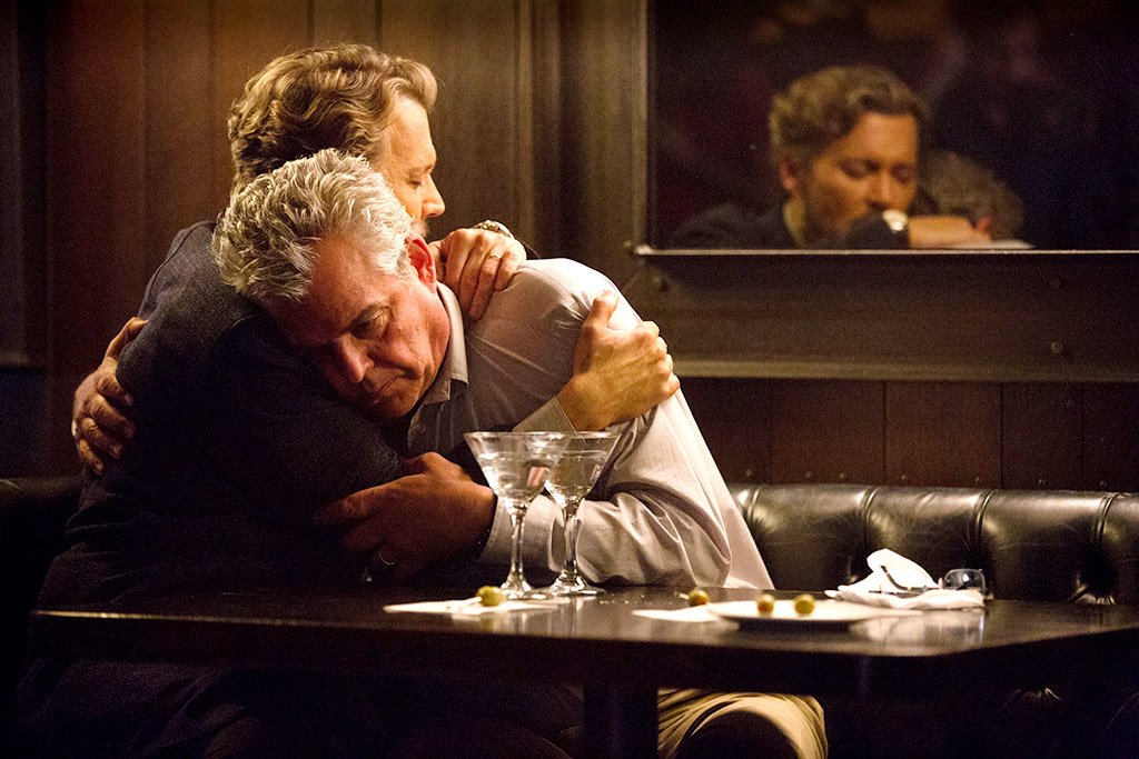 Two older men hugging across a table in a dimly lit restaurant or bar with drinks on the table, showing a moment of comfort or support.