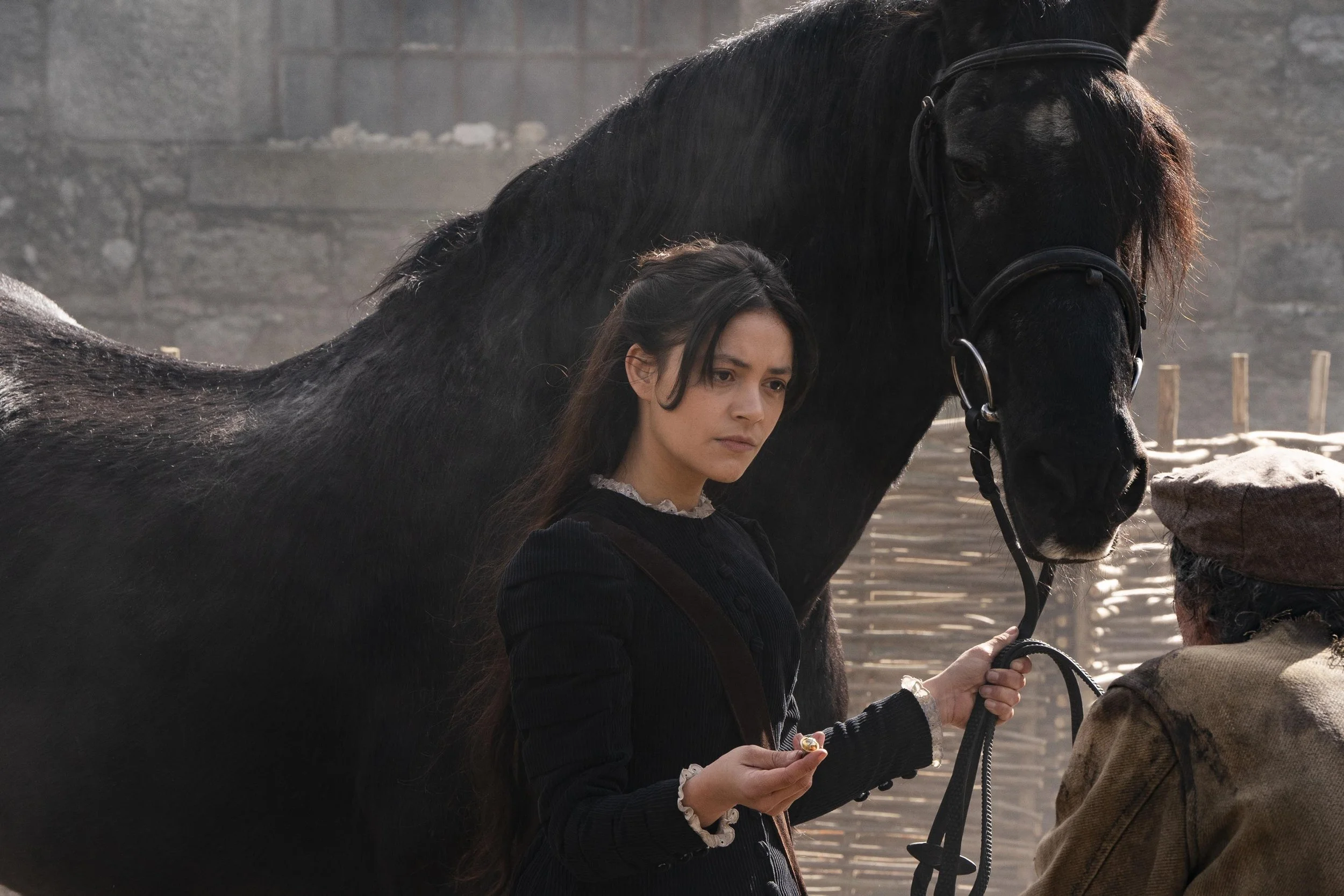 A woman with long dark hair and a black outfit standing next to a black horse. She is holding a riding crop and looking slightly downward, with a serious expression. The background shows a fenced outdoor area with wooden stakes and a brick wall.