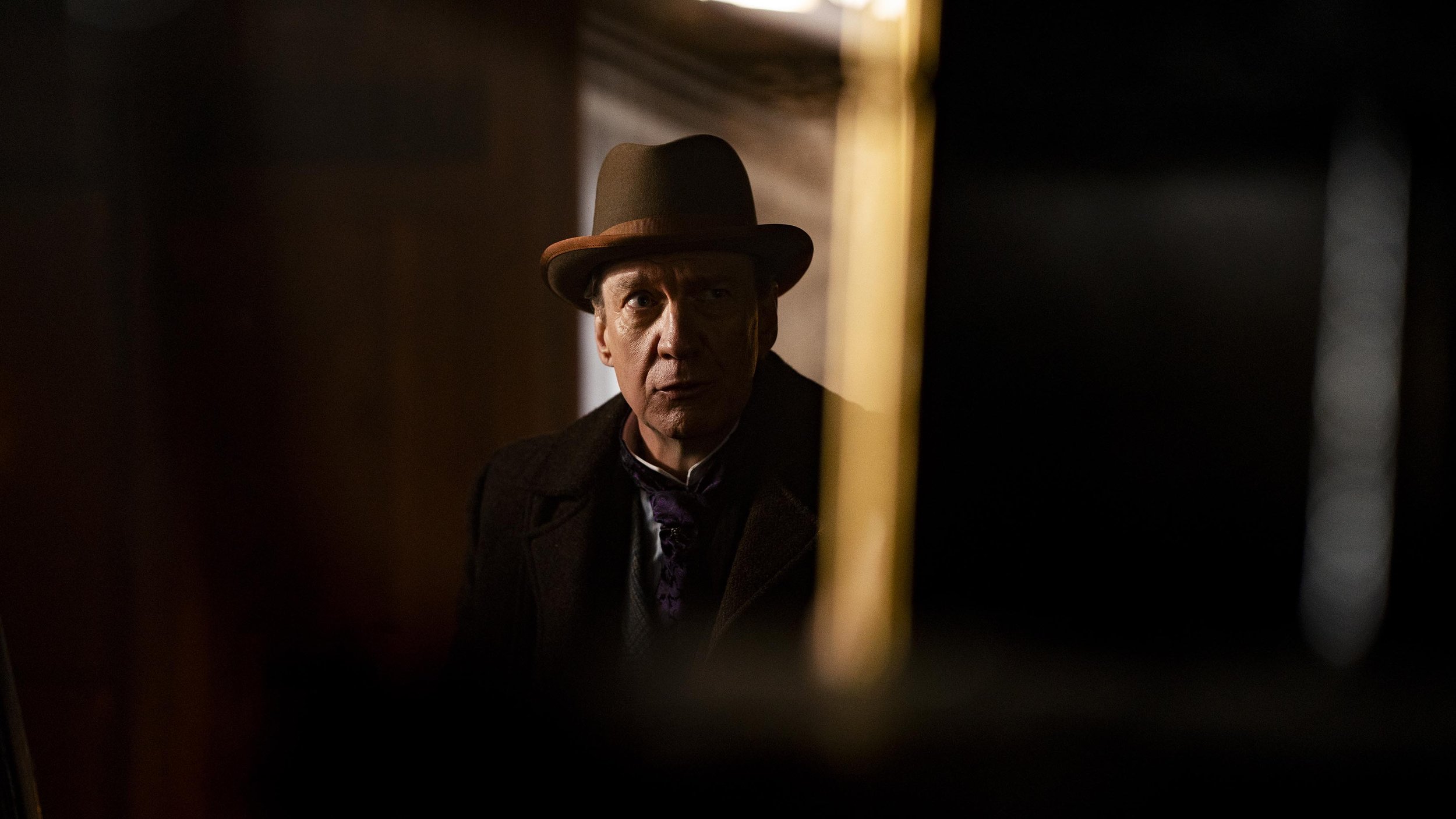 A man wearing a brown fedora hat and dark clothing, looking intently through a narrow opening or gap, with dim lighting and shadowed surroundings.