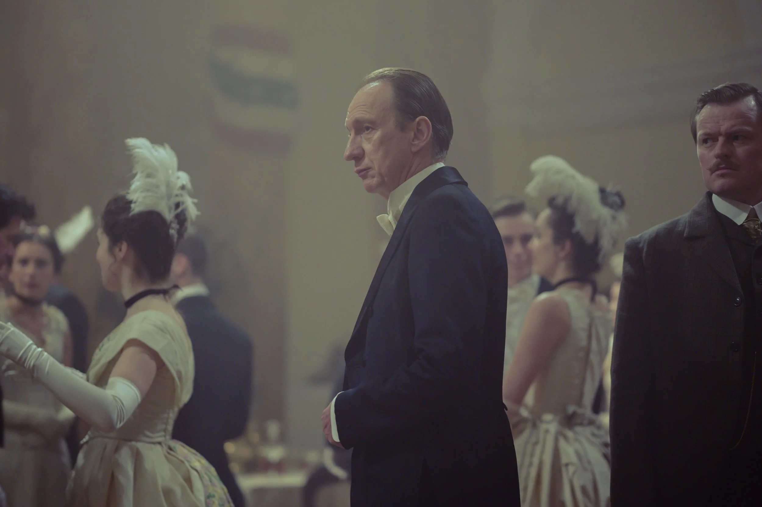 Men and women dressed in 19th-century fancy attire, such as tuxedos and elegant dresses, gather at a formal dance or ball in a grand hall with subdued lighting.