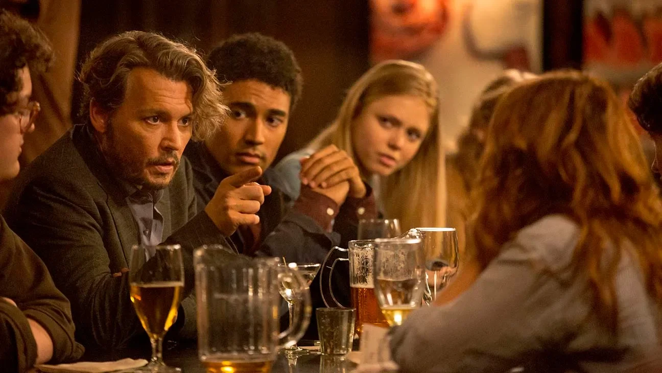 A group of people sitting at a bar, engaged in conversation. There are drinks on the table, including a pitcher of beer, glasses of beer, a glass of white wine, and a glass of red wine. The setting appears to be a cozy, dimly lit bar or restaurant.