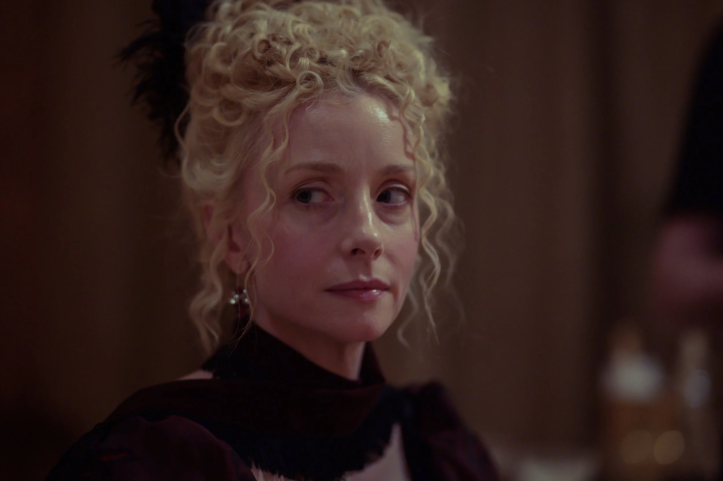 A woman with blonde, curly hair styled in an updo, wearing earrings, and a dark top, looking slightly over her shoulder in a dimly lit setting.