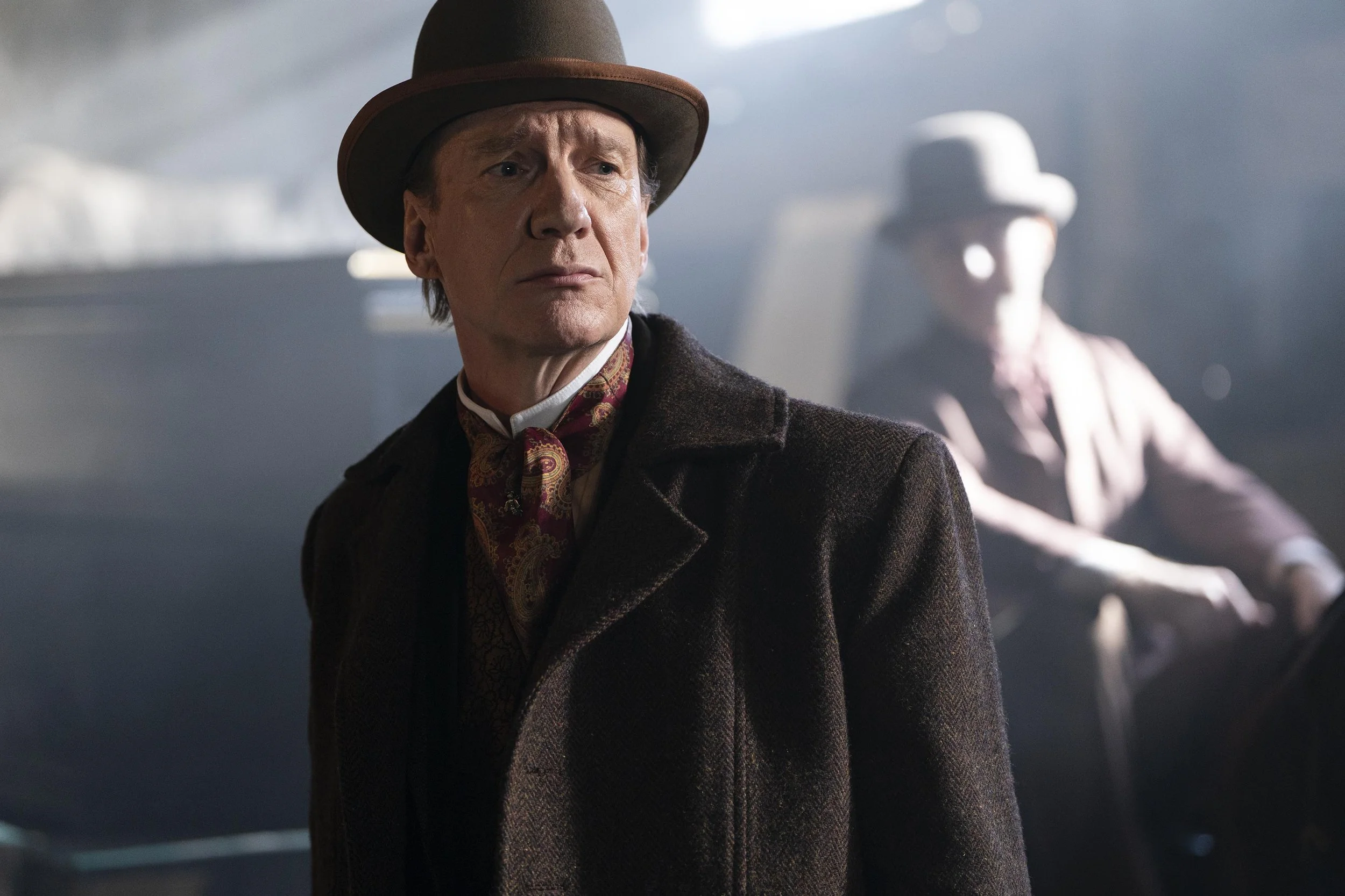 A man dressed in old-fashioned clothing, including a brown suit, a patterned cravat, and a brown hat, standing in a dimly lit room with a contemplative expression, with a woman in a white hat sitting in the background.