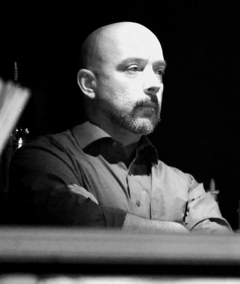 A black and white photo of a man with a beard and a shaved head, wearing a collared shirt, with arms crossed, looking to the side in a serious or contemplative pose.