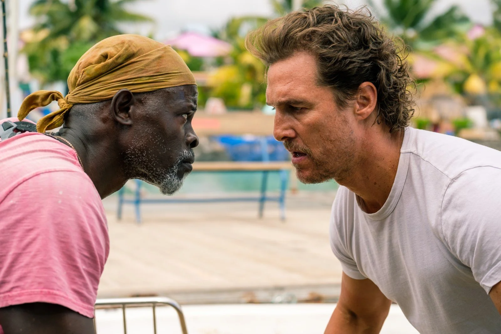 Two men are face to face with intense expressions, leaning in towards each other. One is an older man with a beard, wearing a bandana and a pink shirt. The other is a younger man with curly hair in a white T-shirt. The scene is outdoors, with blurred colorful umbrellas and trees in the background.