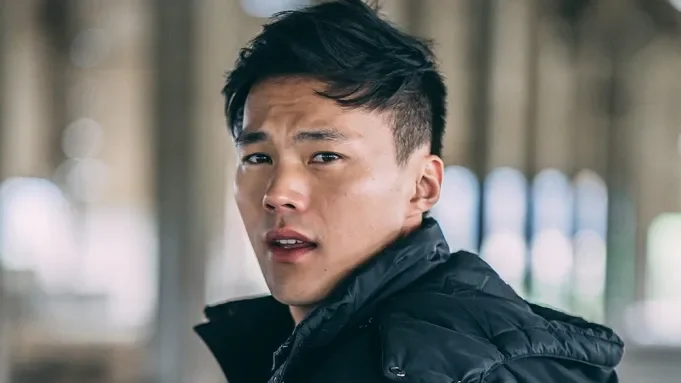 A young man with black hair looking over his shoulder, wearing a black jacket, outdoors with a blurred background of trees and vertical structures.