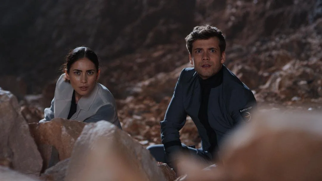 Two people, a woman and a man, crouching behind rocks in a rocky outdoor setting, looking intently ahead, with serious expressions.