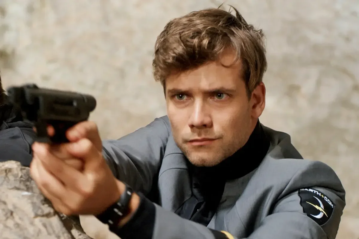 Man aiming a handgun while crouching behind cover, with a serious expression.