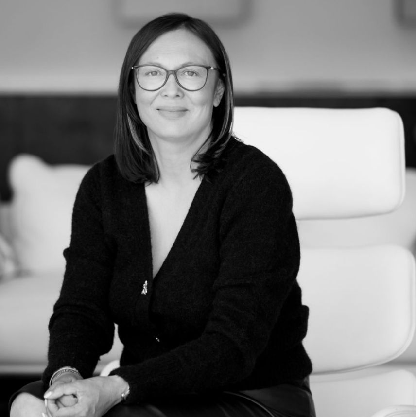 A woman with glasses and shoulder-length hair sits in a modern chair, smiling at the camera, in a black-and-white photo.