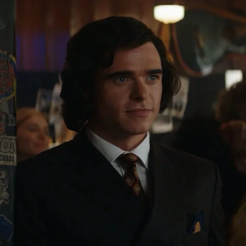 A man with wavy dark hair, wearing a black suit and tie, standing in a dimly lit bar or pub with posters and a neon sign in the background.