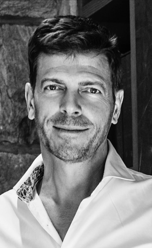 Black and white photo of a smiling man with short, dark hair, wearing a collared shirt, standing outdoors near a stone wall.