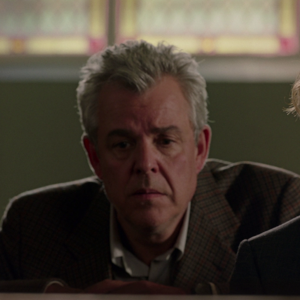 A middle-aged man with gray hair, wearing a checked blazer, sitting indoors with a serious expression on his face, with some colorful images blurred in the background.