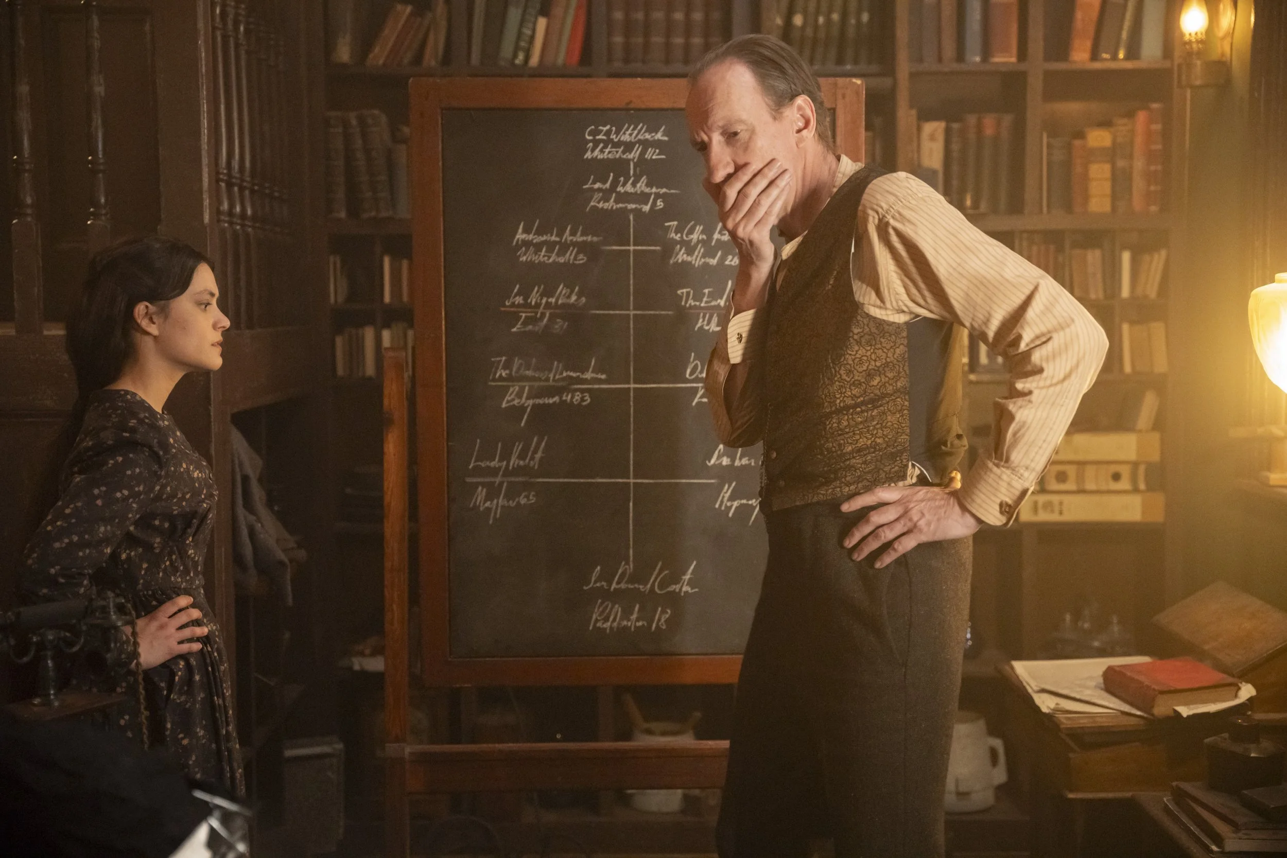 A woman and a man are engaged in a conversation in a study room filled with books. The woman, with dark hair, is standing with her hand on her hip, listening intently. The man, wearing a vest and striped shirt, is leaning forward with his hand on his chin, appearing thoughtful. A chalkboard with writing is visible behind them, along with bookshelves filled with books and a warm light illuminating the scene.