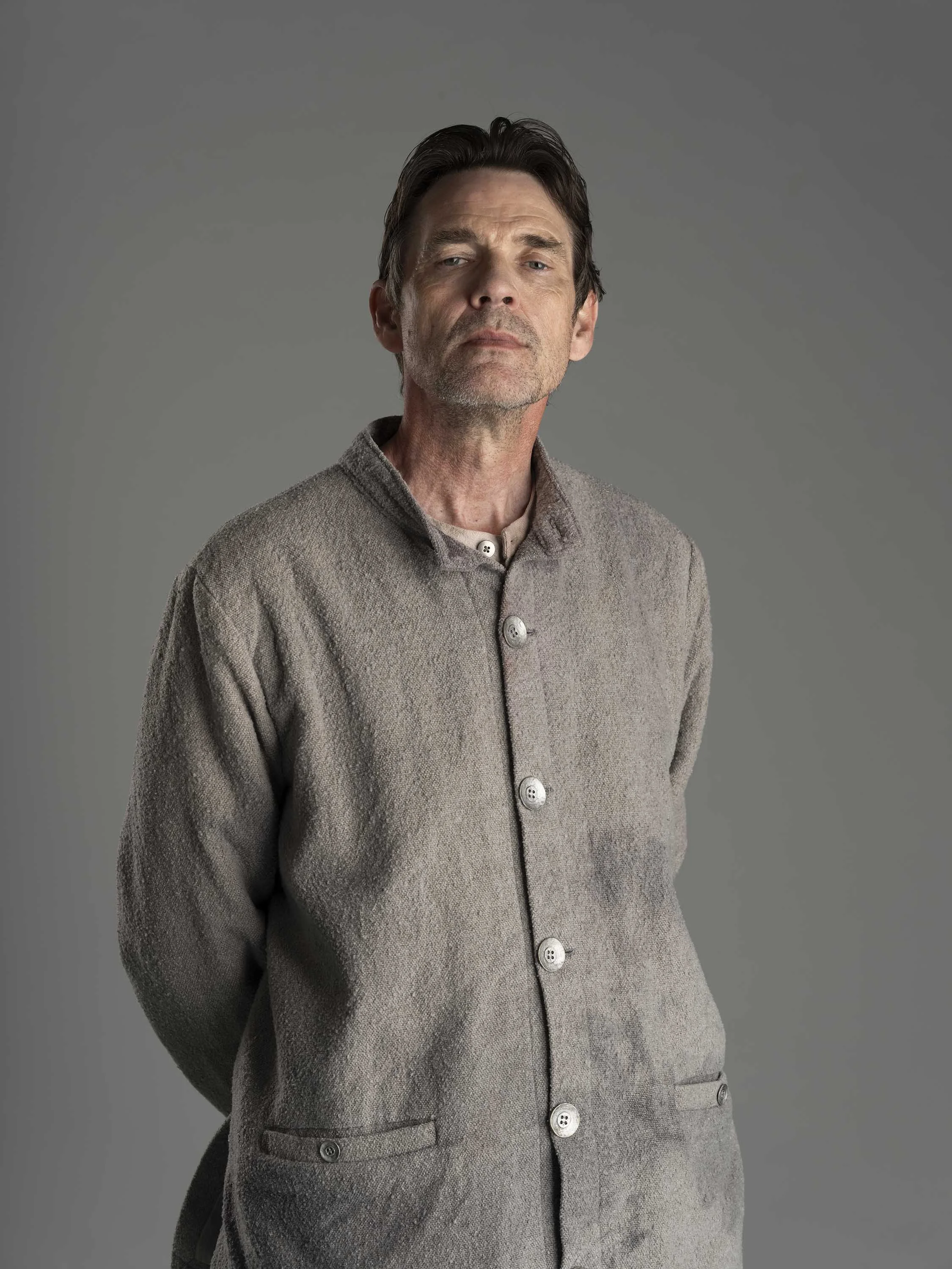 Head and shoulders portrait of a middle-aged man with dark hair, slightly graying, wearing a gray button-up coat with large buttons, standing against a plain gray background.