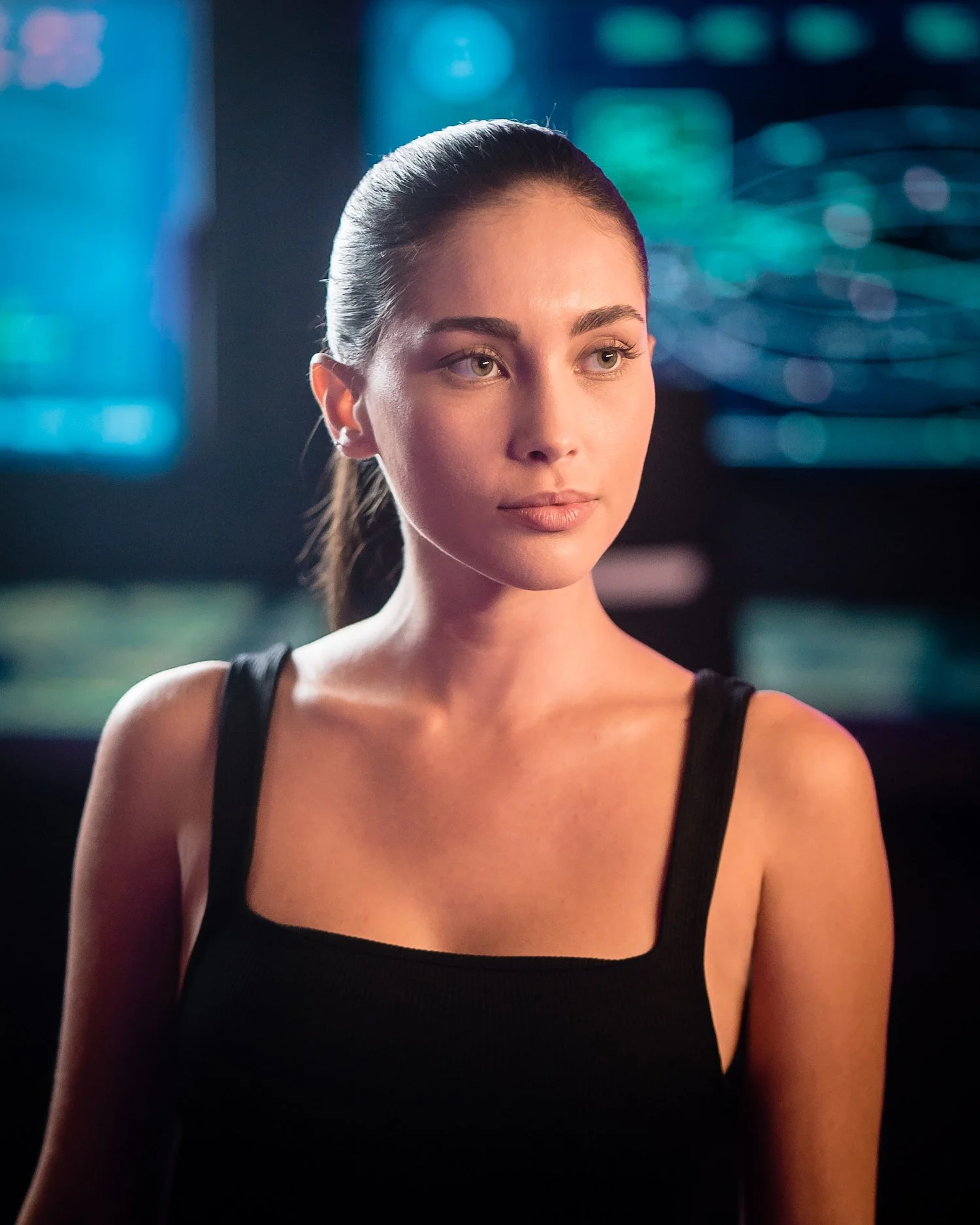 A young woman with dark hair tied back, wearing a black tank top, standing in a dimly lit room with colorful, blurred background lights.