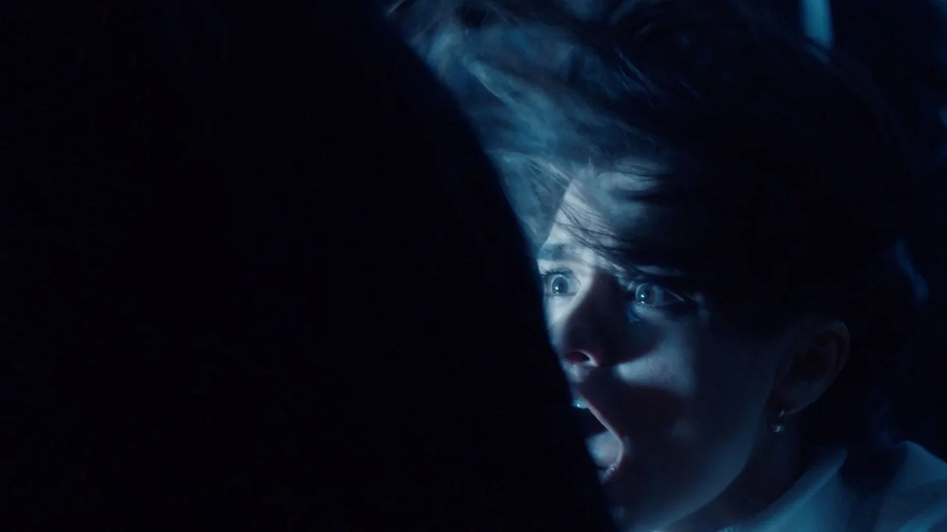Close-up of a person with wide eyes and an open mouth, shadowed in darkness with blue tones, appearing to scream or shout.