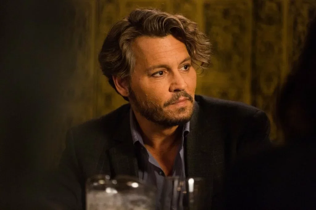 A man with wavy, graying hair and a beard, wearing a dark blazer and shirt, sitting in a dimly lit setting.