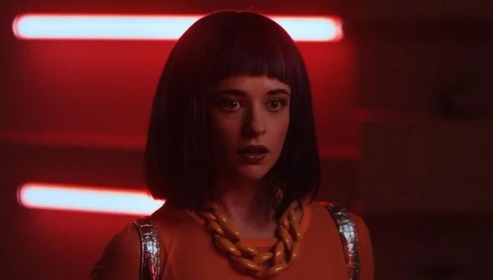 Woman with short dark hair, wearing an orange top and chunky yellow necklace, standing in front of red neon lights.