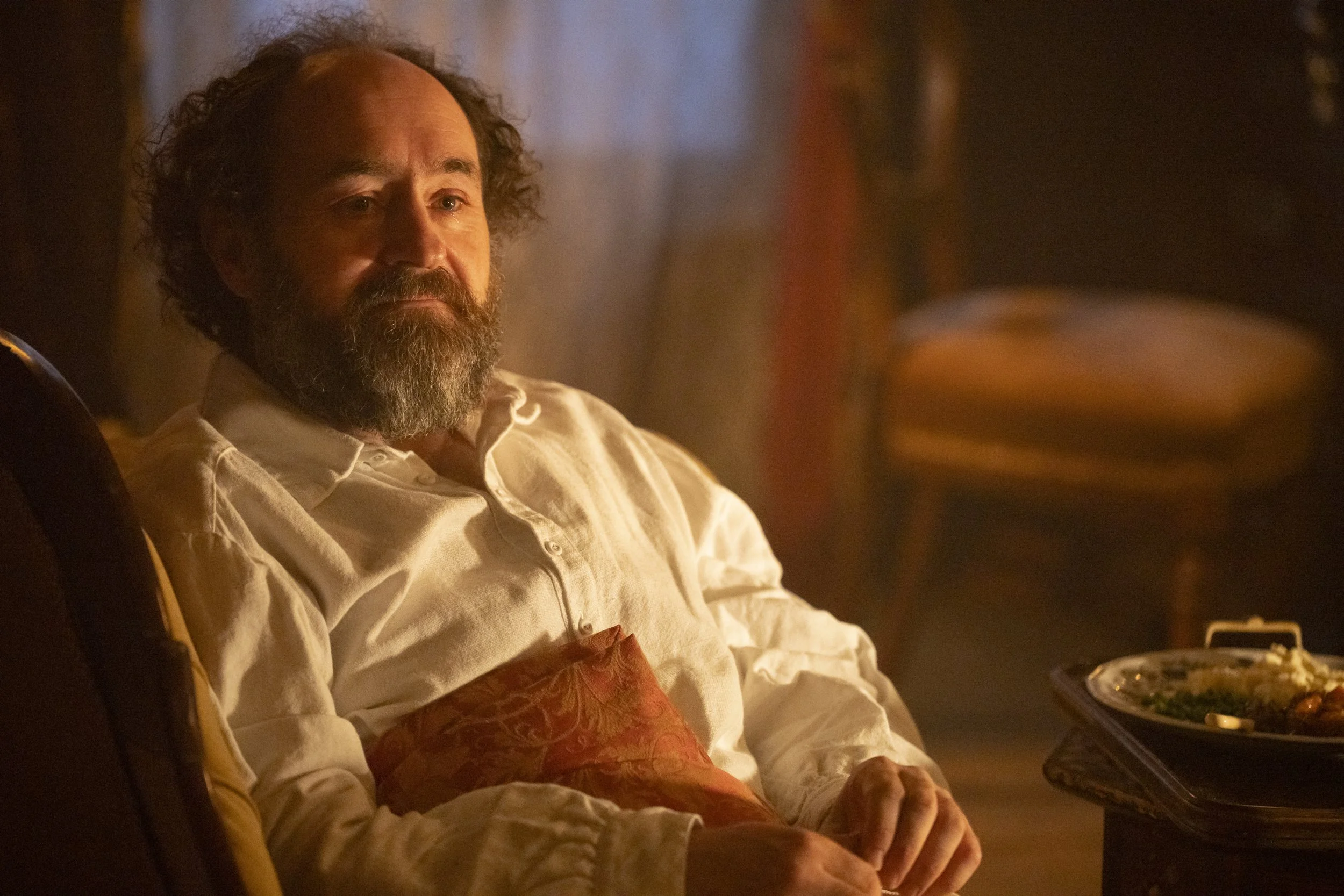 A man with curly hair and a beard sitting at a dining table in a dimly lit room.