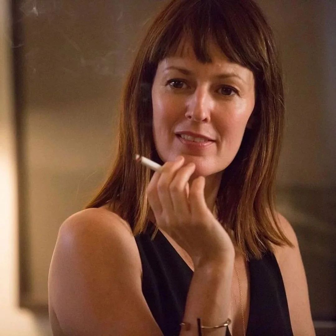 A woman with shoulder-length brown hair, wearing a black sleeveless top, holds a cigarette near her face and looks to the side in a serious expression.