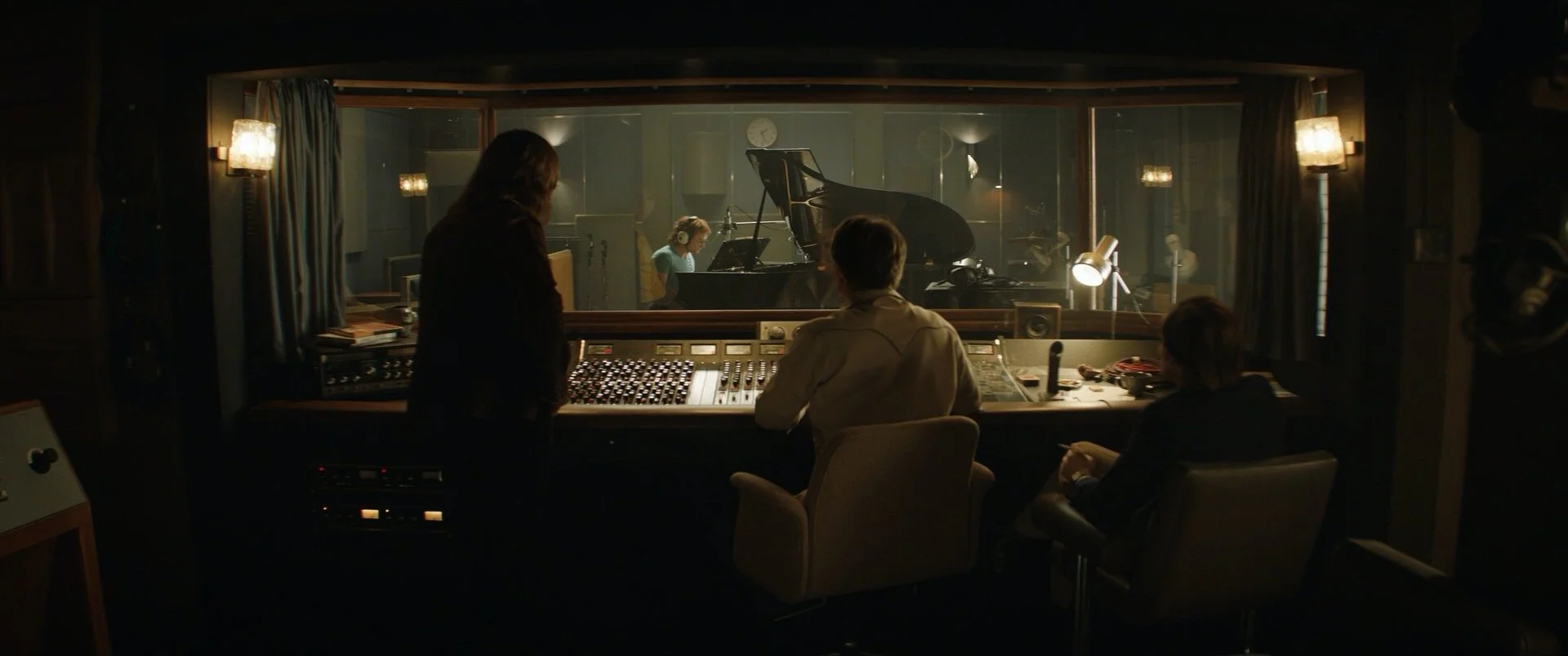 Recording studio with three people in control room, one woman and two men sitting at mixing console, woman standing, looking into glass window to musicians playing piano and guitar in studio.
