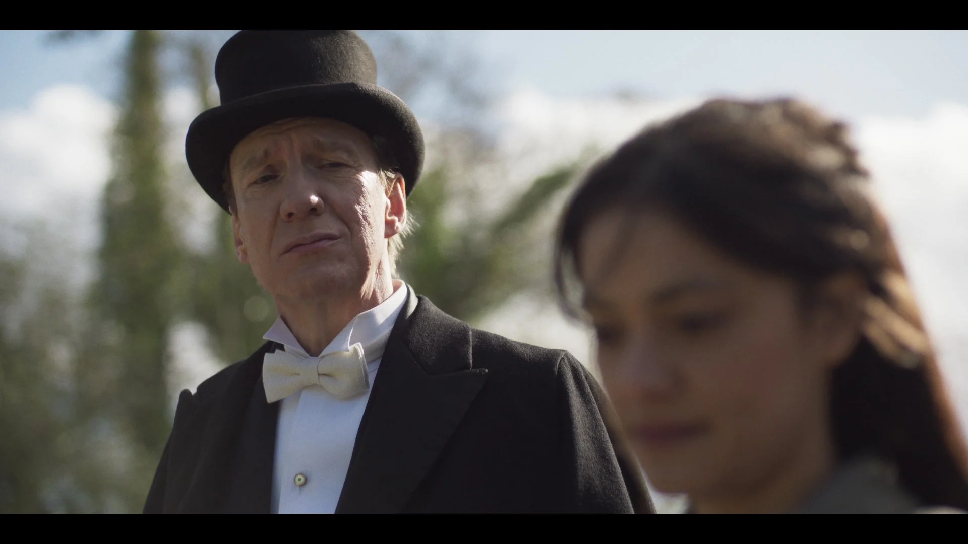 A man dressed as Abraham Lincoln, wearing a black top hat, white bowtie, and black tuxedo, looks down at a woman with dark hair. The scene is outdoors with blurred trees and sky in the background.