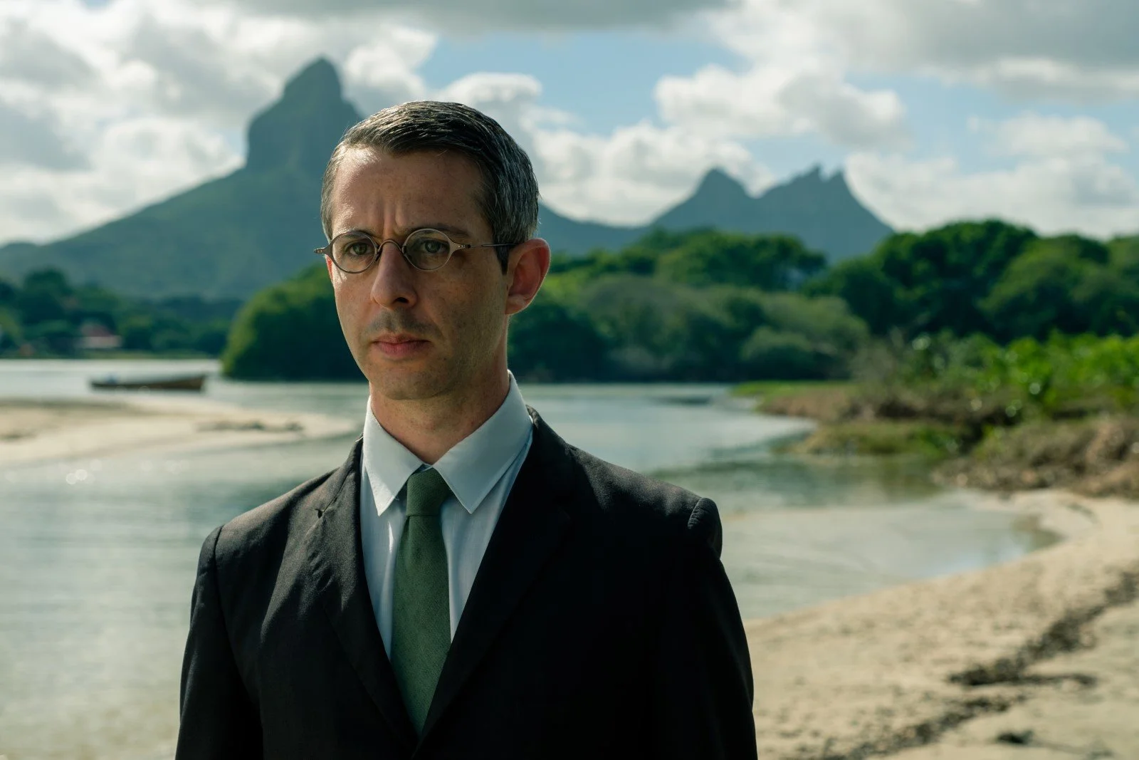 A man in a black suit and green tie standing outdoors near a river with mountains and lush green trees in the background.