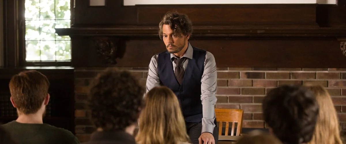 A man with brown wavy hair and a beard stands in front of a small group, in a room with brick walls and a window, giving a lecture or presentation.