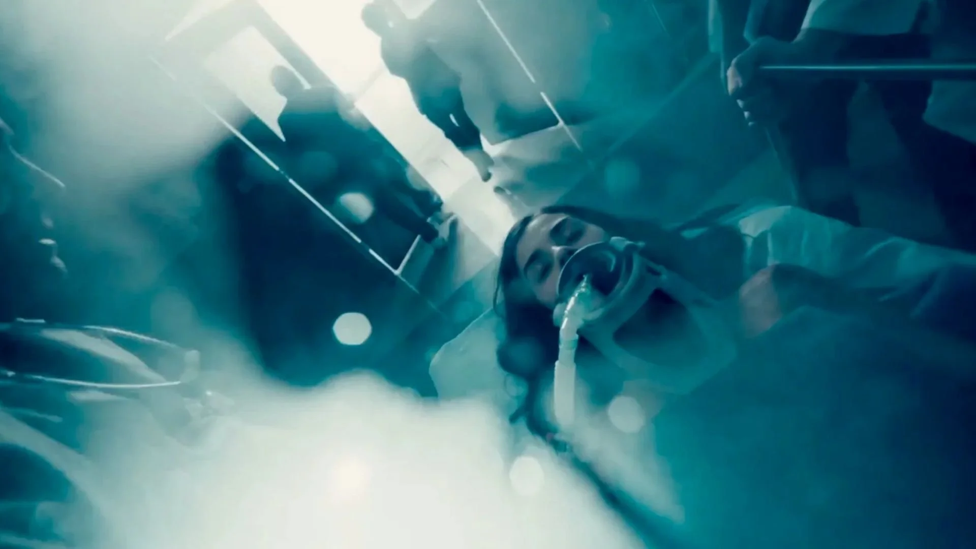 A scene from a sci-fi or action movie, showing a person in a spacesuit or armored suit lying on the ground, with a breathing mask connected to a tube, amid fog or smoke and blue lighting.