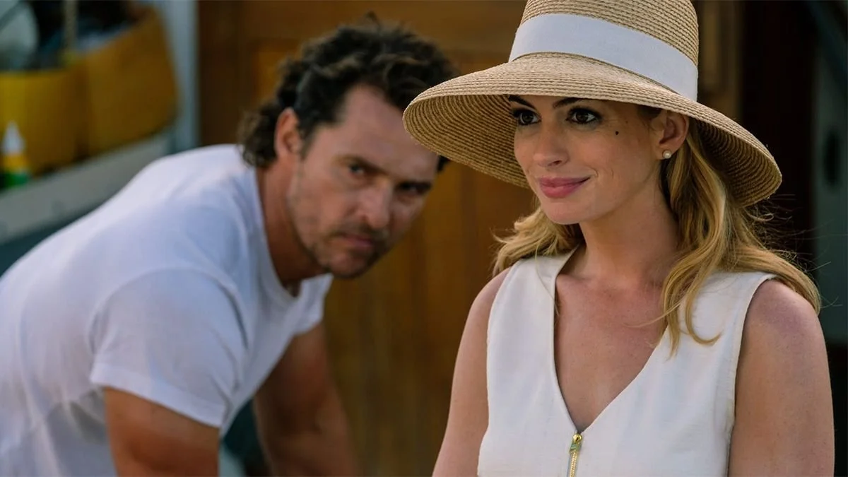 A woman wearing a large sun hat and sleeveless top, smiling, with a man in the background leaning forward.