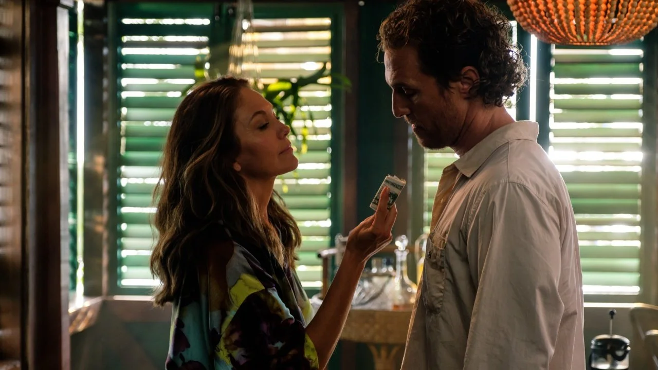 A woman holding money in front of a man in a dimly lit room with green shutters in the background.