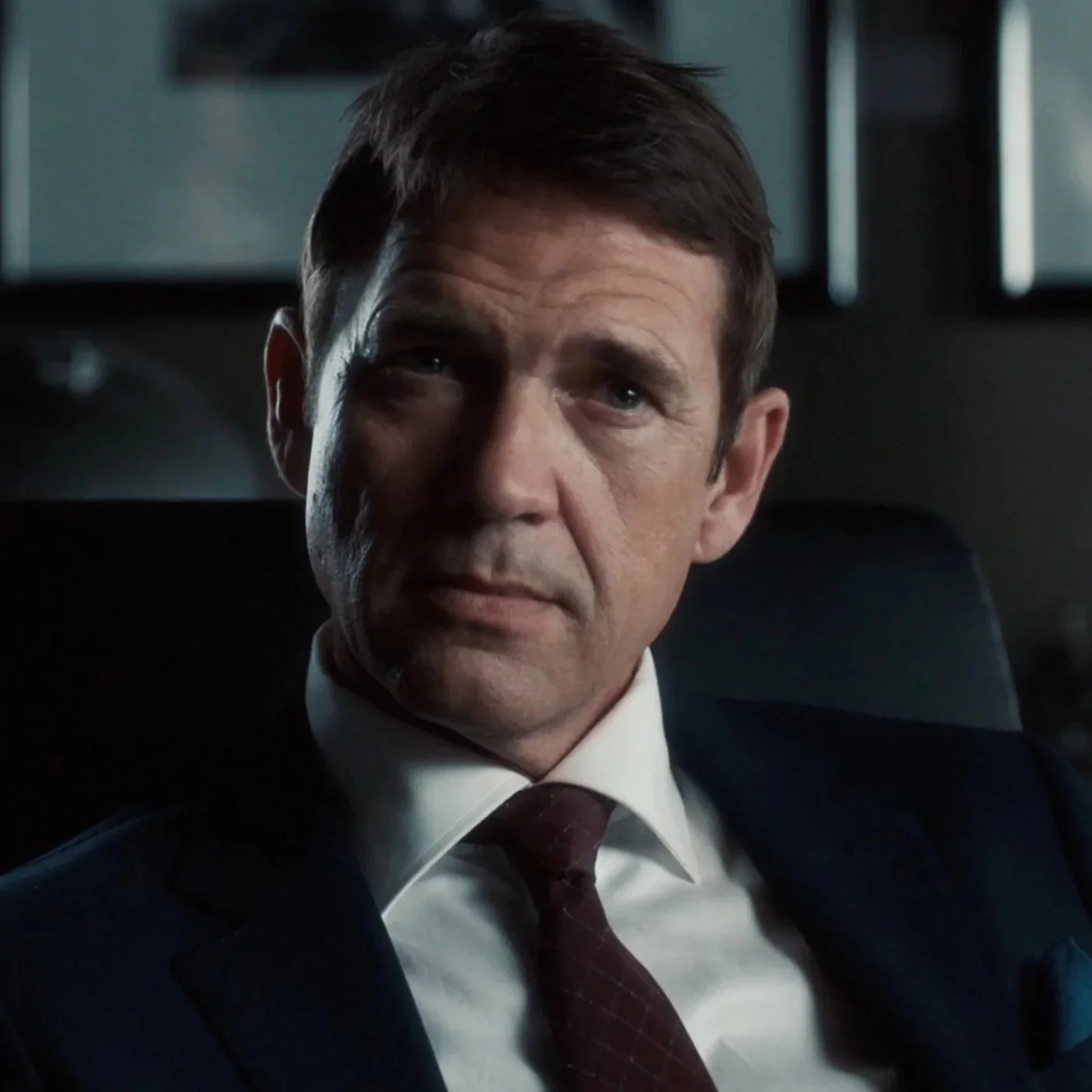 A middle-aged man with short brown hair, wearing a dark suit, white shirt, and maroon tie, sitting inside a vehicle.