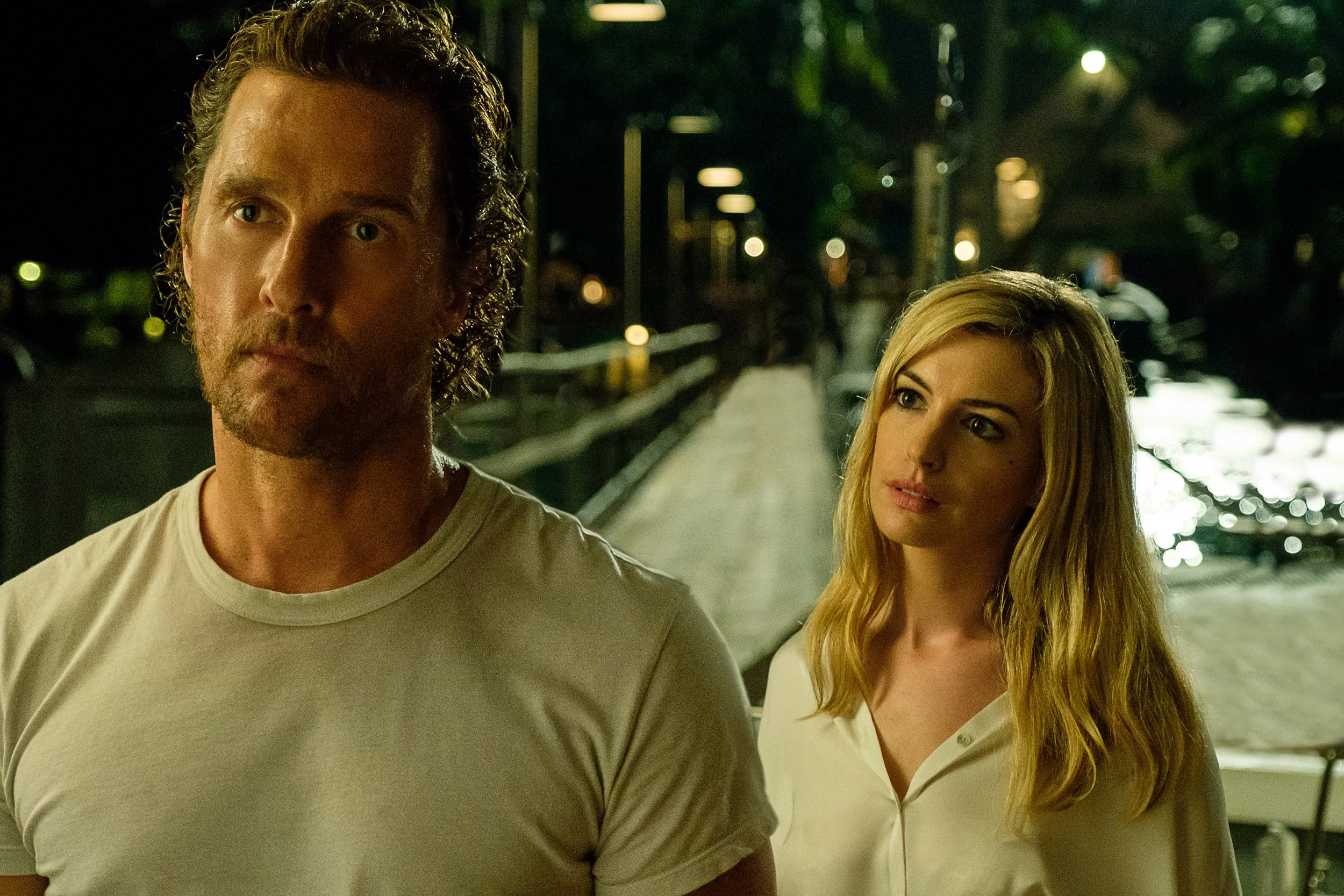 A man and a woman walking at night near a river or canal with streetlights in the background. The man has curly brown hair, a beard, and is wearing a light-colored t-shirt. The woman has long blonde hair, fair skin, and is wearing a white blouse.
