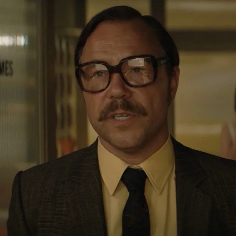 Man with dark hair, mustache, glasses, wearing a dark suit with a yellow shirt and dark tie, indoors in a professional setting.
