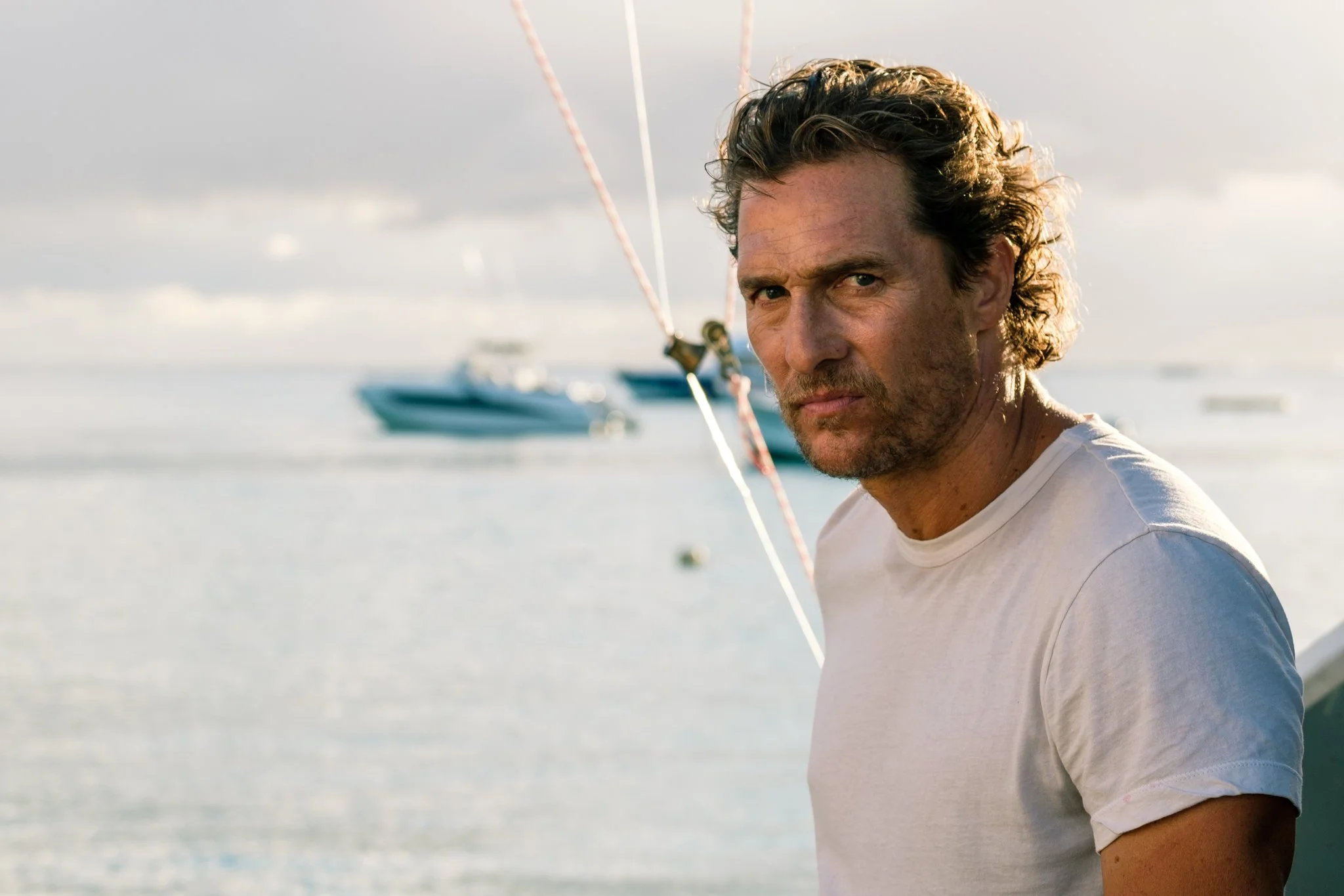 A man with curly hair and a beard standing outdoors near water with boats in the background.