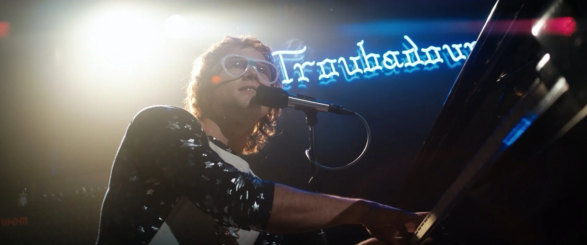 A man with curly hair and glasses is playing a piano and singing into a microphone on stage with a neon blue sign in the background.