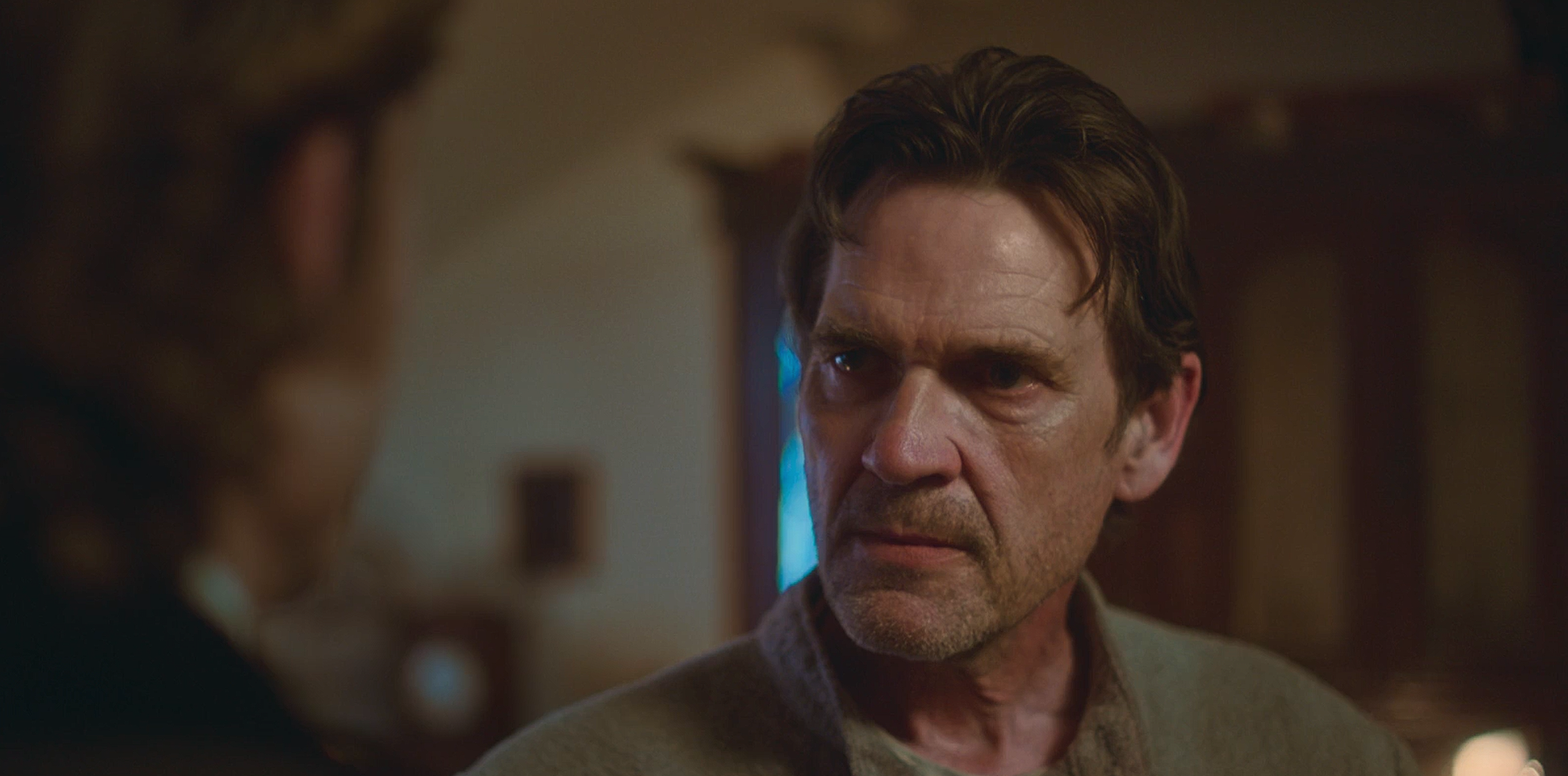 A man with dark hair and stubble looking intensely at another person in a dimly lit room.