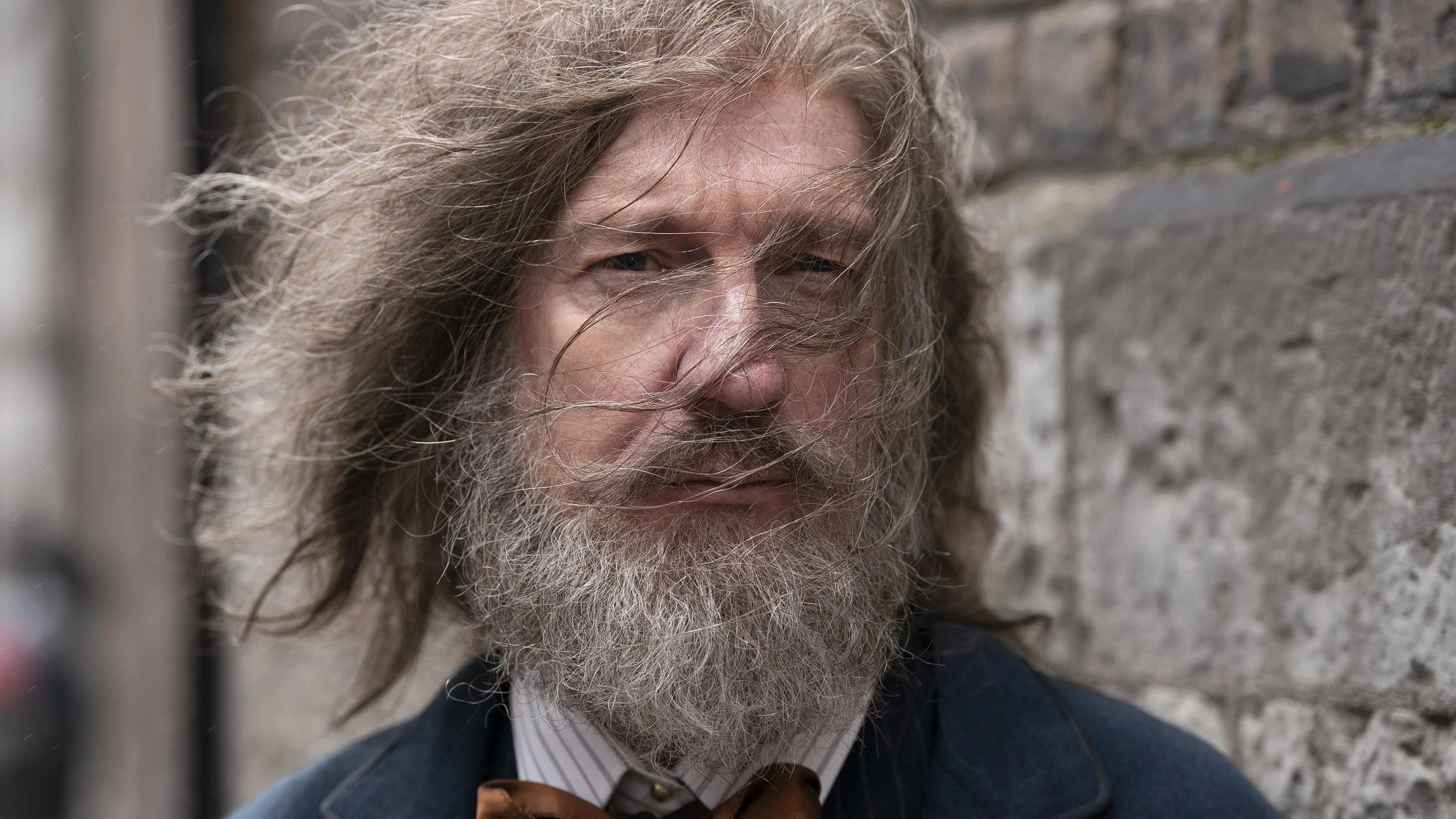 A man with long, curly gray hair and a full beard standing against a stone wall, looking at the camera with a serious expression.