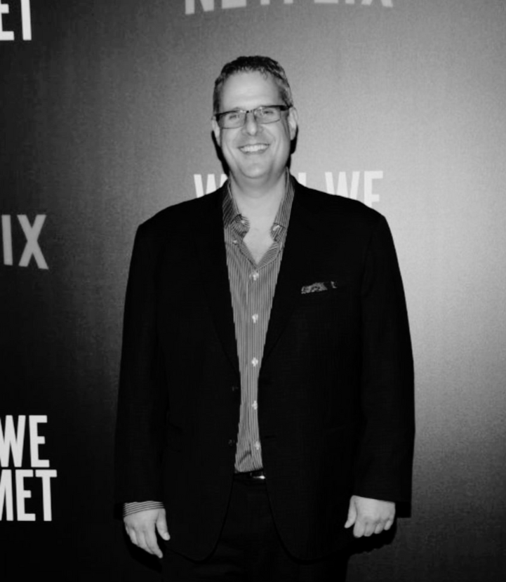 A smiling man with glasses, wearing a suit and striped shirt, standing in front of a backdrop with the Netflix logo and text.