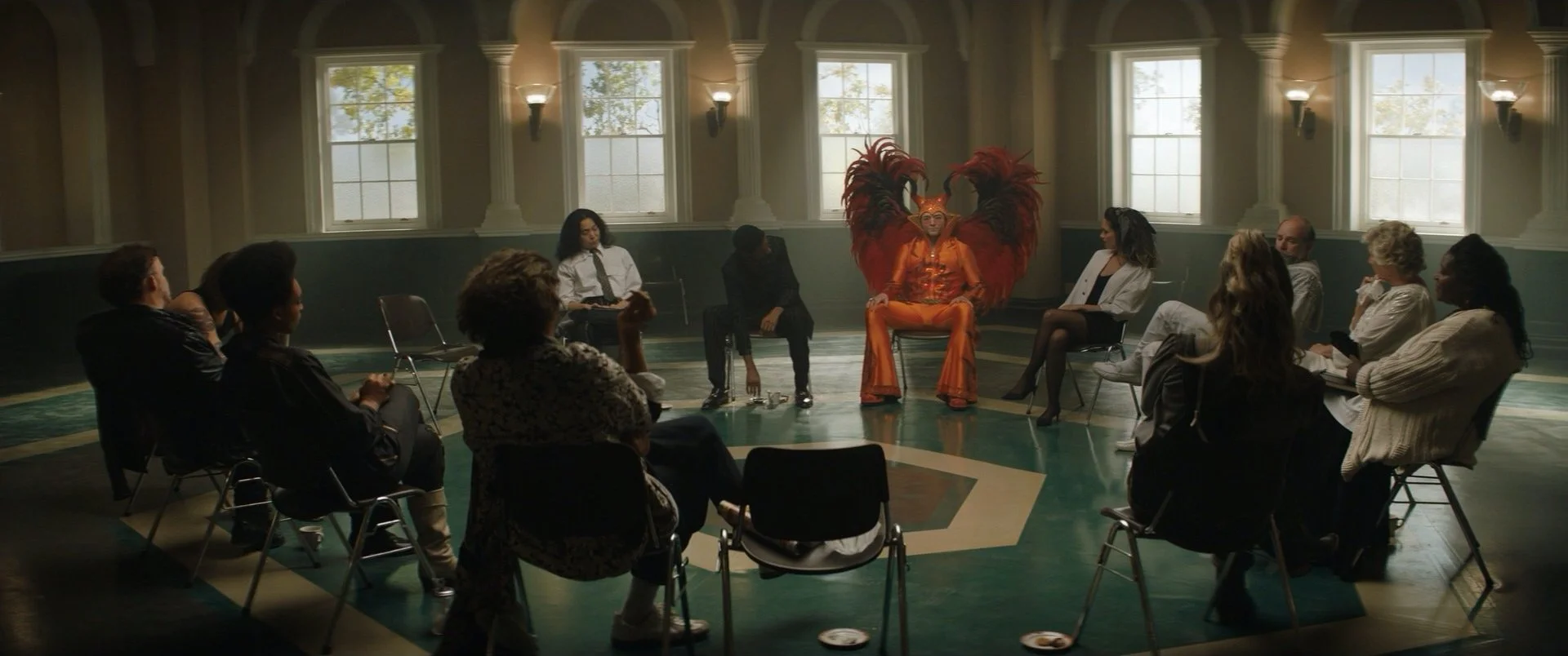 A diverse group of people seated in a circle in a large room with tall windows, with a performer in an elaborate orange costume and feathered headdress in the center.