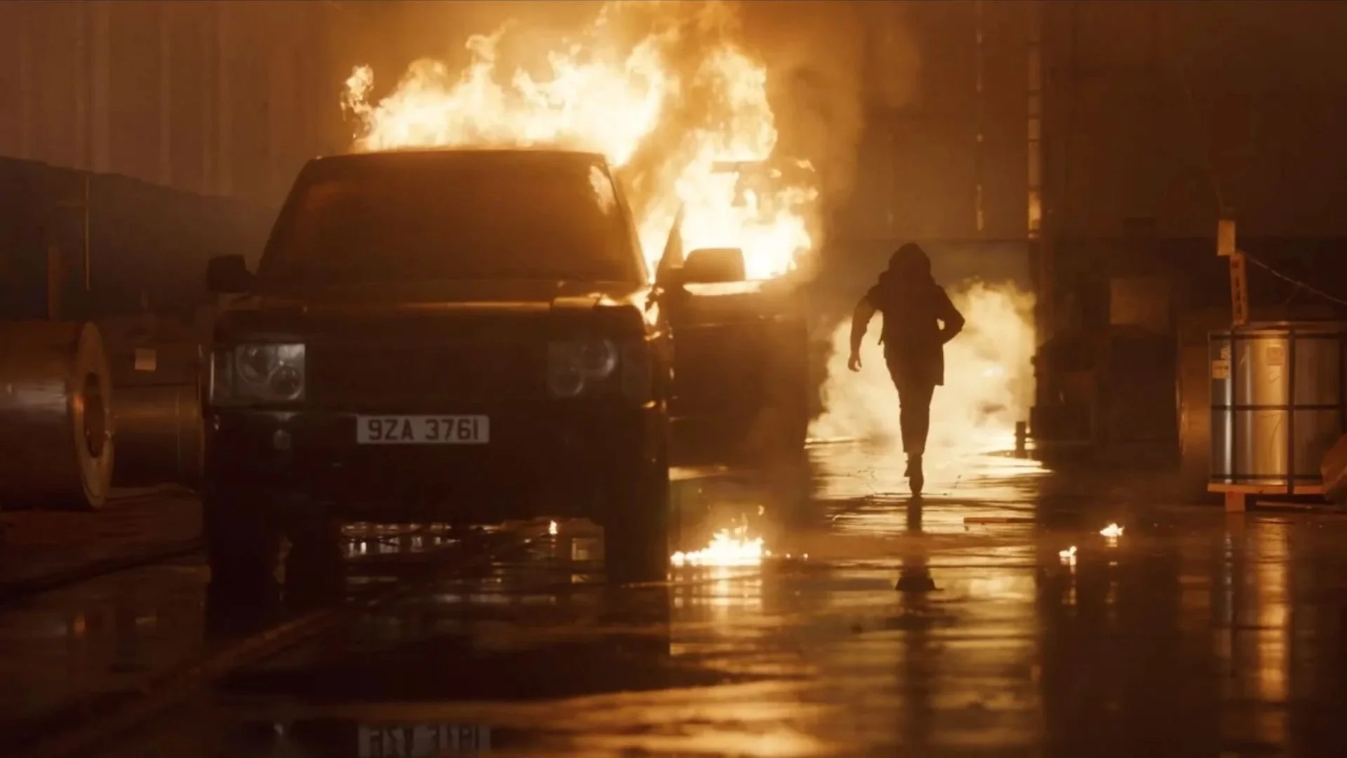 A person walking away from a burning vehicle in a dark, rainy street at night.