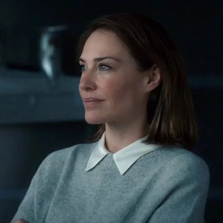 A woman with shoulder-length brown hair wearing a light gray sweater with a white collared shirt underneath, sitting in a dimly lit room.
