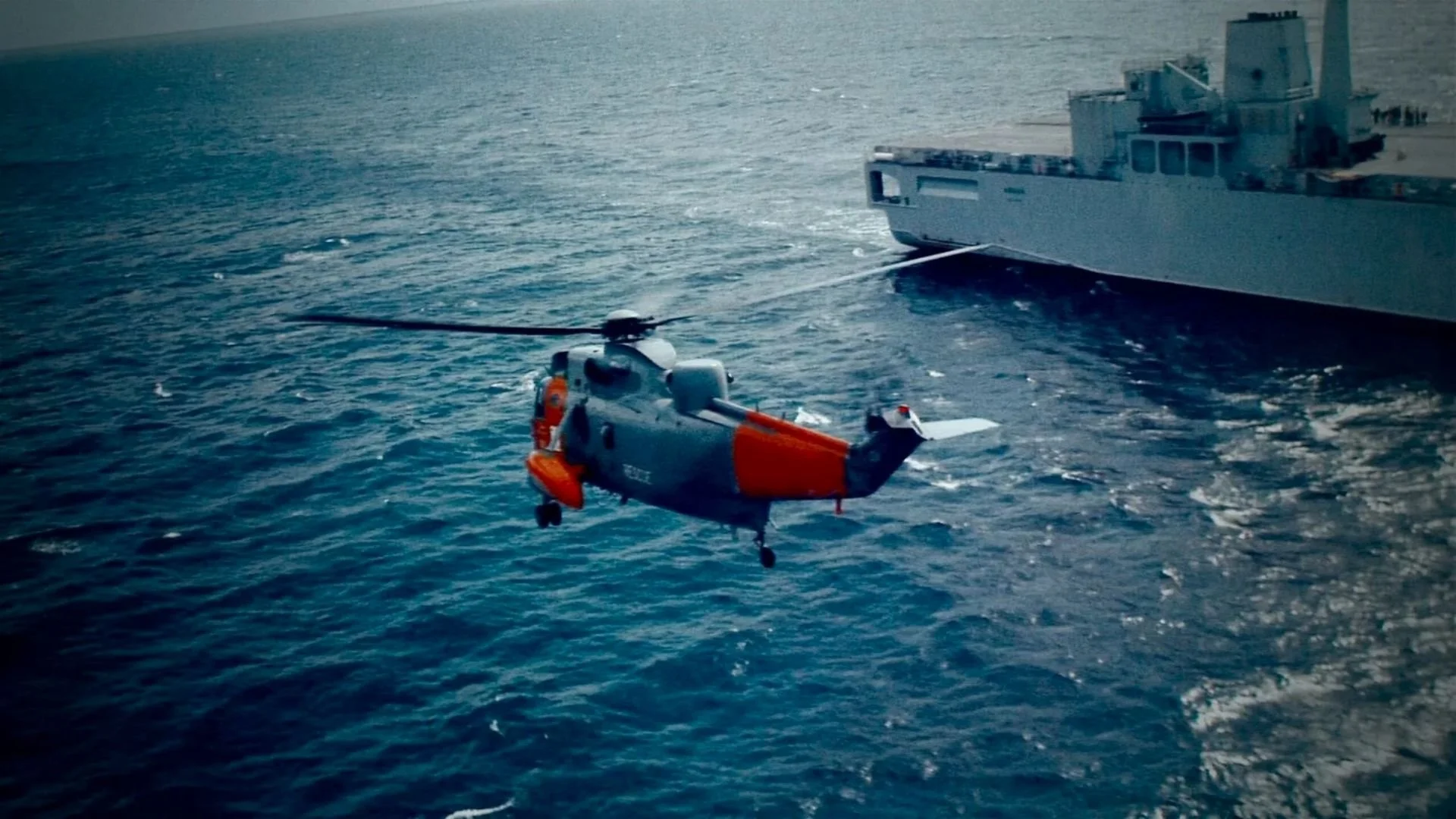 A helicopter flying over the ocean near a large gray ship.
