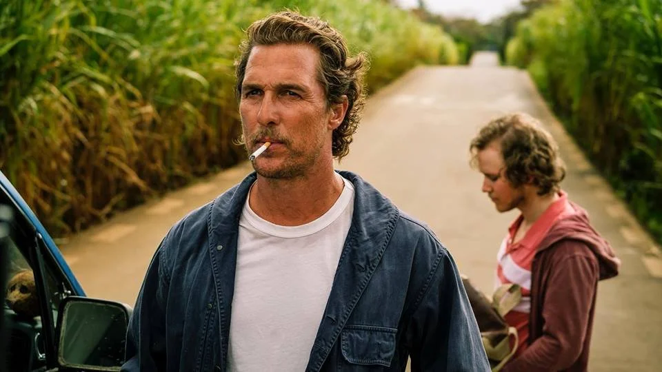 A man with curly hair, wearing a white t-shirt and a dark jacket, stands outdoors on a rural road, smoking a cigarette. In the background, a woman with curly hair, glasses, and a pink hoodie looks down and interacts with an animal.