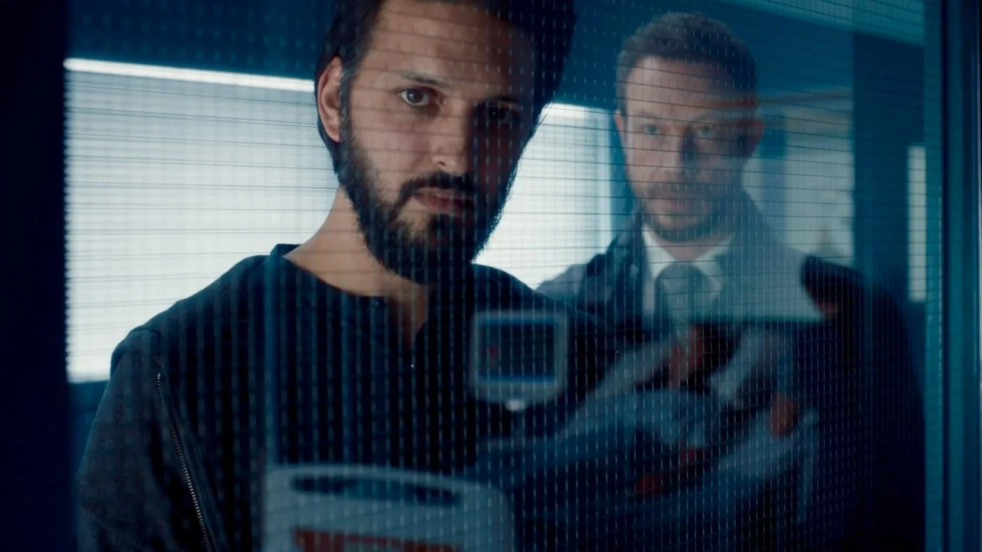 Two men are looking through a glass window, with one man in the foreground and the other in the background. The man in the foreground has dark hair and a beard, and is holding a smartphone. The man in the background has short hair and is wearing a suit with a tie.