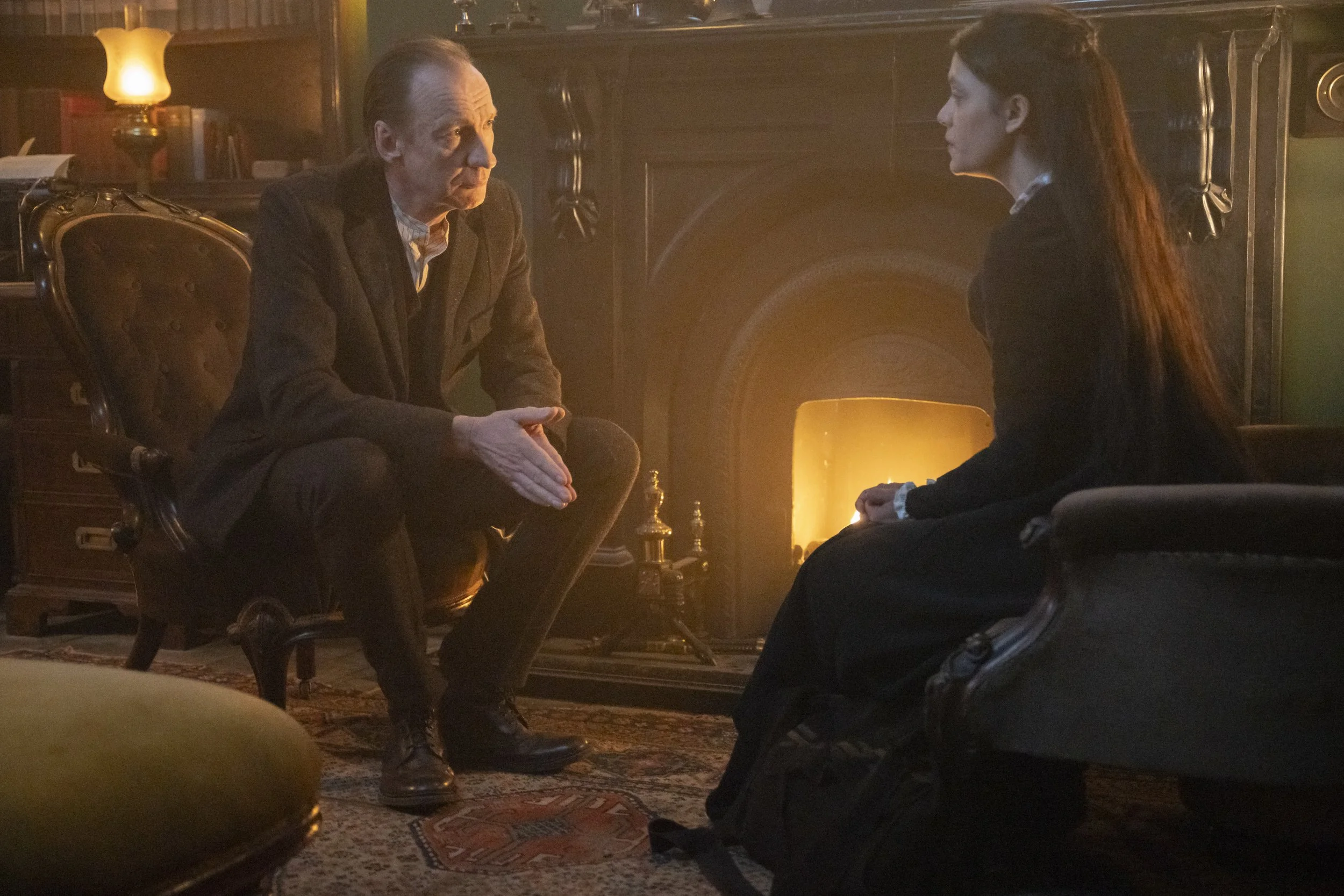Two people sitting in a dimly lit room having a serious conversation. An older man with light skin and slicked-back hair is sitting on the left, facing a woman with light skin and long, reddish hair sitting on the right. They are seated across from each other near a fireplace with a warm glow, surrounded by books and antique furniture.