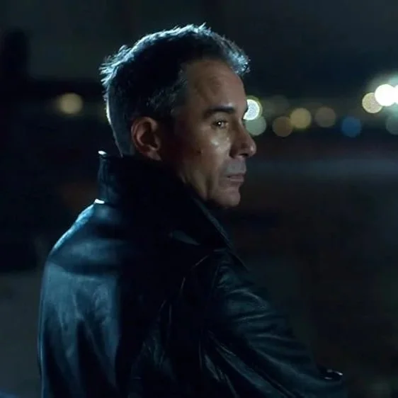 A man with dark hair and a leather jacket looking over his shoulder at night with blurred city lights in the background.