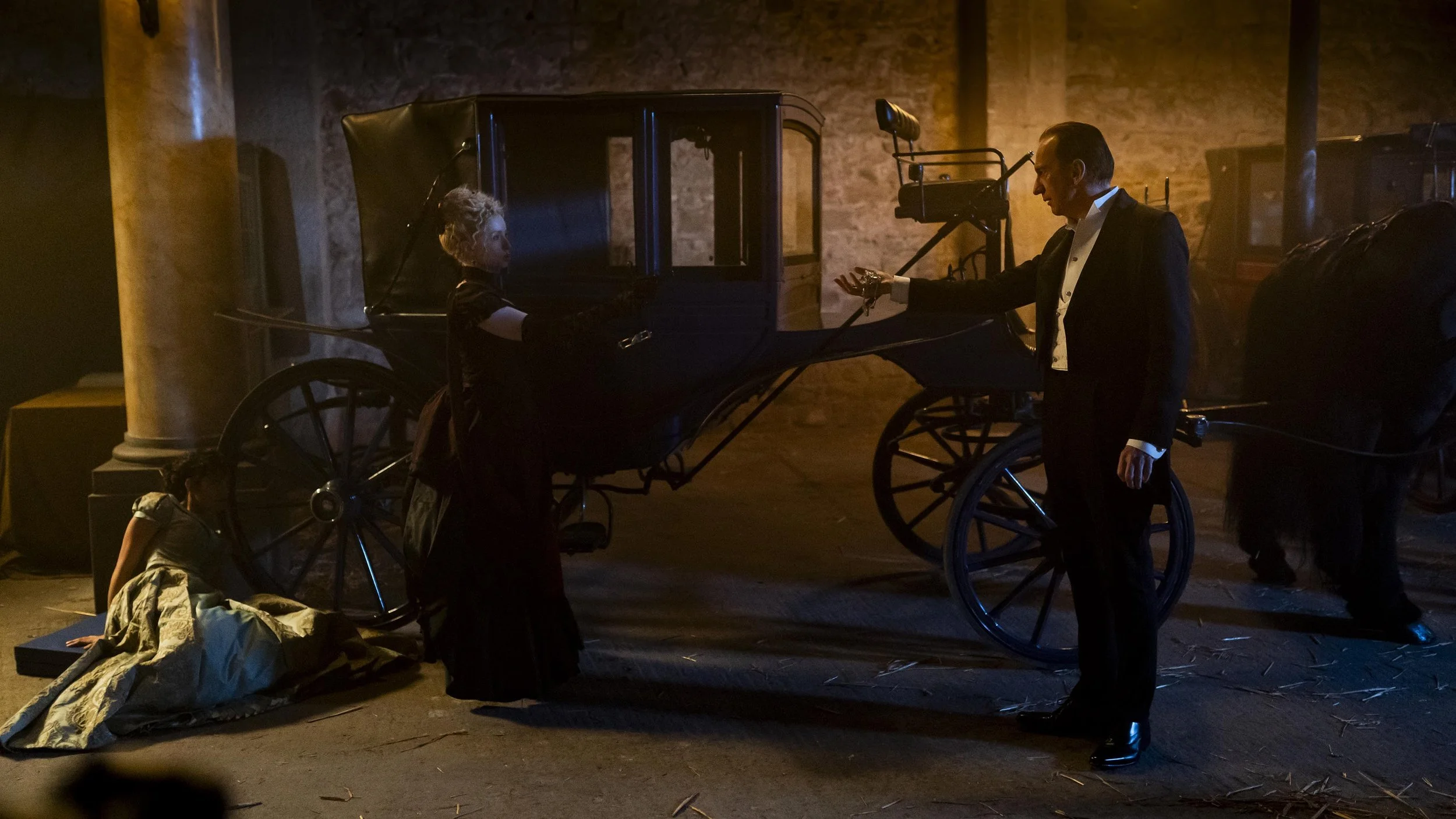 A scene from a theatrical performance showing a man in a tuxedo and a woman in a dress standing by a vintage horse-drawn carriage, with children in costumes sitting or lying on the floor, set against a brick wall background with dramatic lighting.